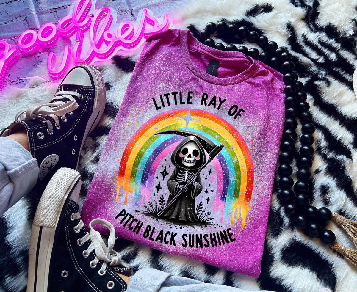 Close-up of bleach-wash tee showing dripping rainbow over a skeleton reaper holding a scythe with “Little Ray Of Pitch Black Sunshine” text.
