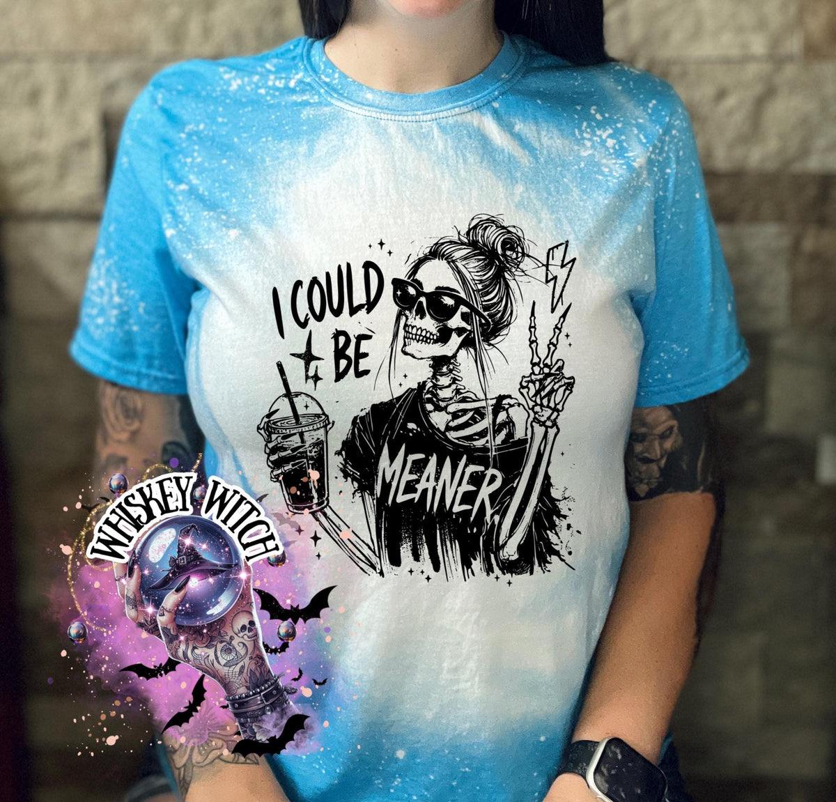 A saphire blue  t-shirt with bleach white splatter effect with a graphic of a skeleton wearing sunglasses, holding a drink, and making a peace sign. The text on the shirt reads "I COULD BE MEANER.”