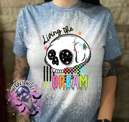 Hand-bleached indigo blue  graphic tee featuring a skull illustration, small stars, and the words “Living the Dream” with rainbow drip lettering.