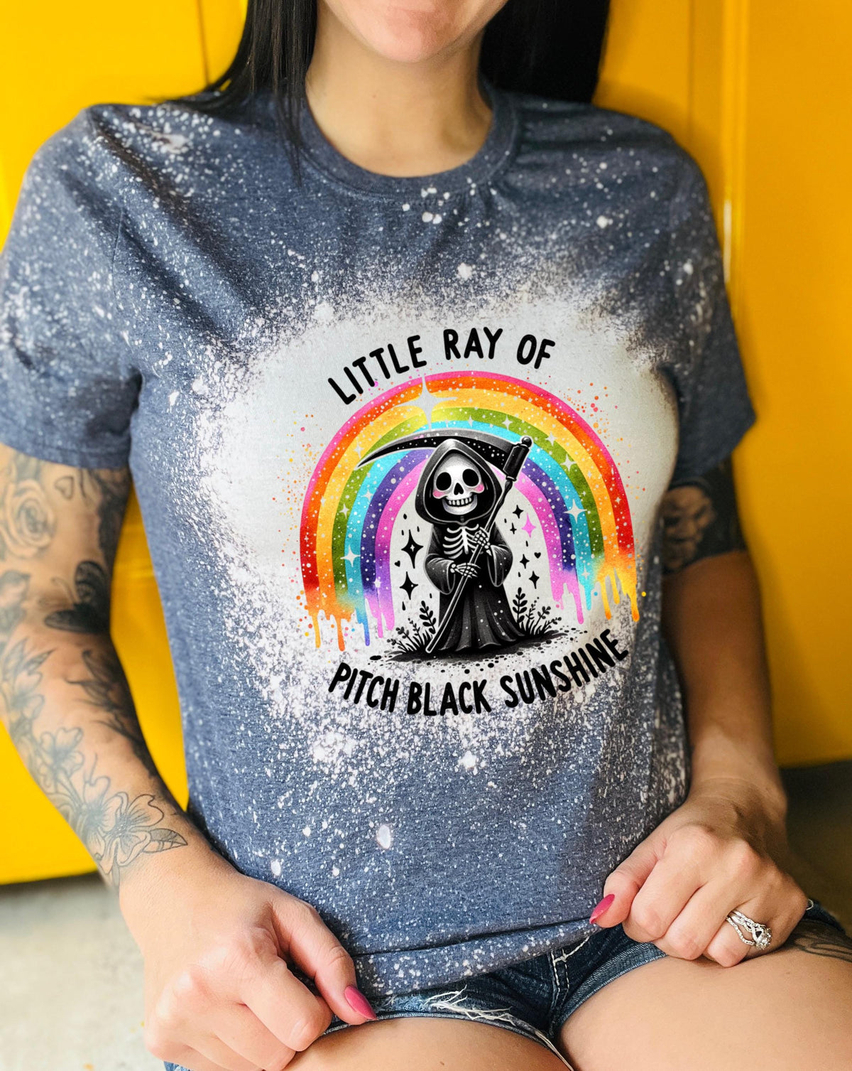 Close-up of bleach-wash tee showing dripping rainbow over a skeleton reaper holding a scythe with “Little Ray Of Pitch Black Sunshine” text.
