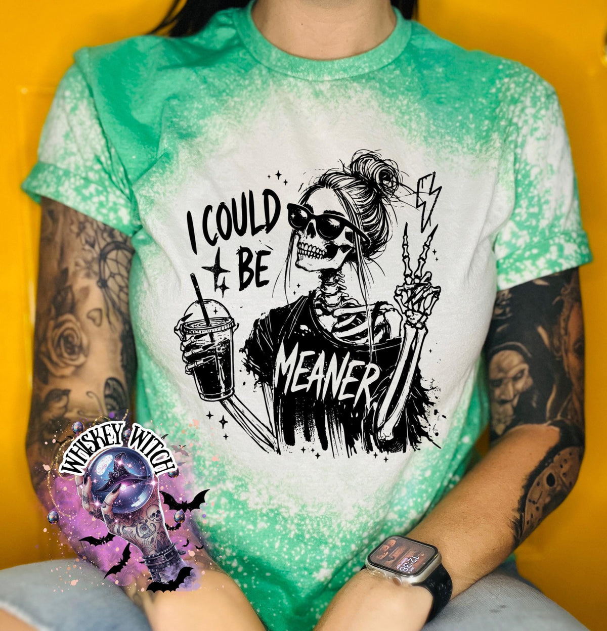 A green t-shirt with bleach white splatter effect with a graphic of a skeleton wearing sunglasses, holding a drink, and making a peace sign. The text on the shirt reads "I COULD BE MEANER.”