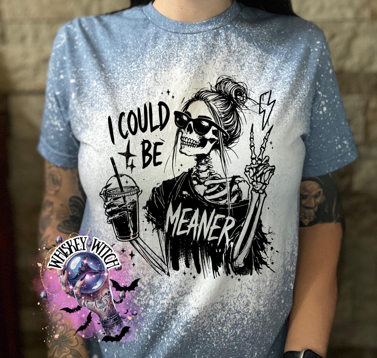 A indigo blue  t-shirt with bleach white splatter effect with a graphic of a skeleton wearing sunglasses, holding a drink, and making a peace sign. The text on the shirt reads "I COULD BE MEANER.”