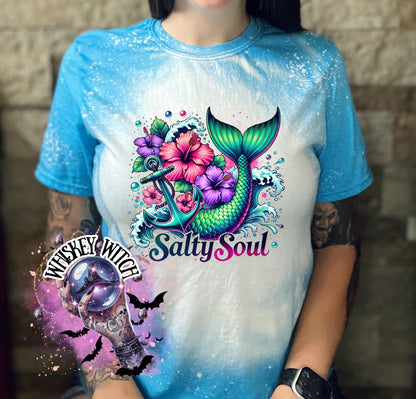 A sapphire blue   t-shirt with a bleach splatter effect. The shirt features a colorful graphic of a mermaid tail, anchor, flowers, and the text "Salty Soul". The mermaid tail is green and blue.