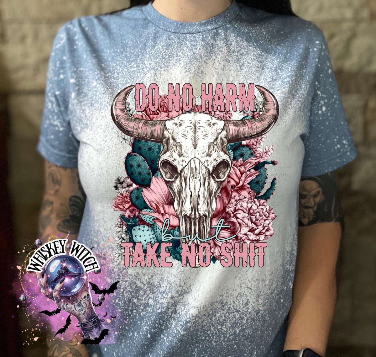 Light blue t-shirt with a bleached effect, featuring a cow skull graphic with pink horns, surrounded by pink flowers and cacti. The text "DO NO HARM" arches above the skull, with "but TAKE NO SHIT" below.