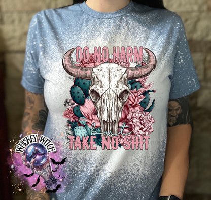 Light blue t-shirt with a bleached effect, featuring a cow skull graphic with pink horns, surrounded by pink flowers and cacti. The text "DO NO HARM" arches above the skull, with "but TAKE NO SHIT" below.