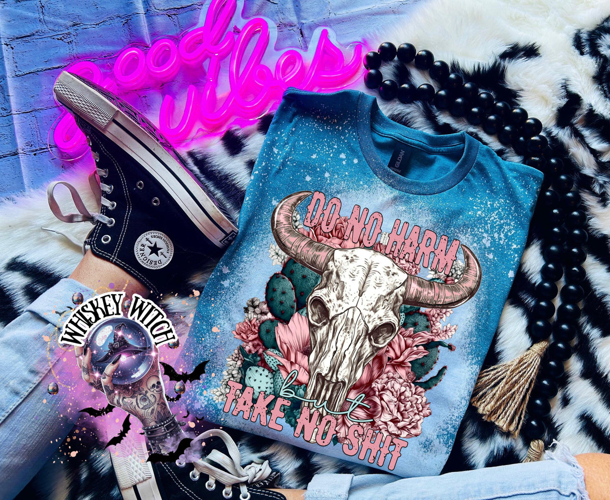 Galapagos blue t-shirt with a bleached effect, featuring a cow skull graphic with pink horns, surrounded by pink flowers and cacti. The text "DO NO HARM" arches above the skull, with "but TAKE NO SHIT" below.