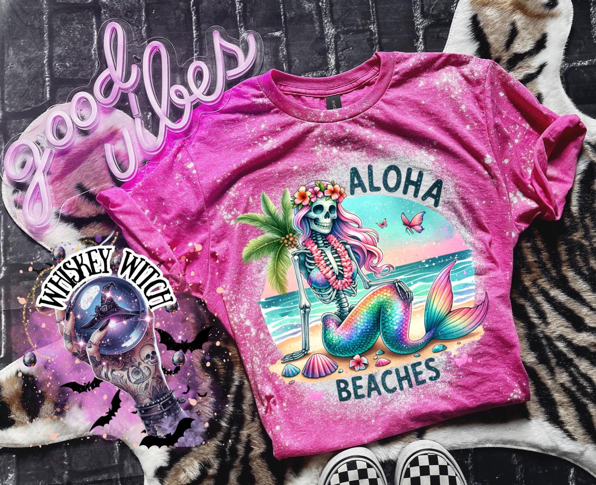 A pink t-shirt with a bleached effect features a colorful skeleton mermaid with a flower crown, palm tree, and seashells. The text "Aloha Beaches" is printed on the shirt.