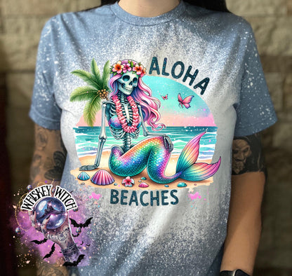 A indigo  blue  t-shirt with a bleached effect features a colorful skeleton mermaid with a flower crown, palm tree, and seashells. The text "Aloha Beaches" is printed on the shirt.