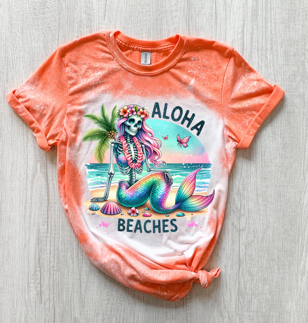 A orange t-shirt with a bleached effect features a colorful skeleton mermaid with a flower crown, palm tree, and seashells. The text "Aloha Beaches" is printed on the shirt.