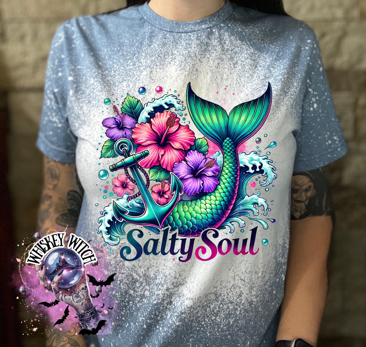 A indigo blue   t-shirt with a bleach splatter effect. The shirt features a colorful graphic of a mermaid tail, anchor, flowers, and the text "Salty Soul". The mermaid tail is green and blue.