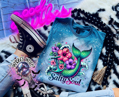 A Galapagos blue  t-shirt with a bleach splatter effect. The shirt features a colorful graphic of a mermaid tail, anchor, flowers, and the text "Salty Soul". The mermaid tail is green and blue.