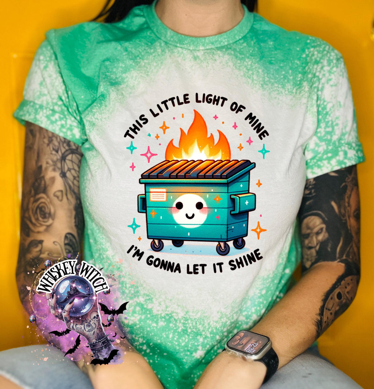green t-shirt with a bleach-splattered design featuring a cartoon dumpster on fire with the text "This little light of mine I'm gonna let it shine".