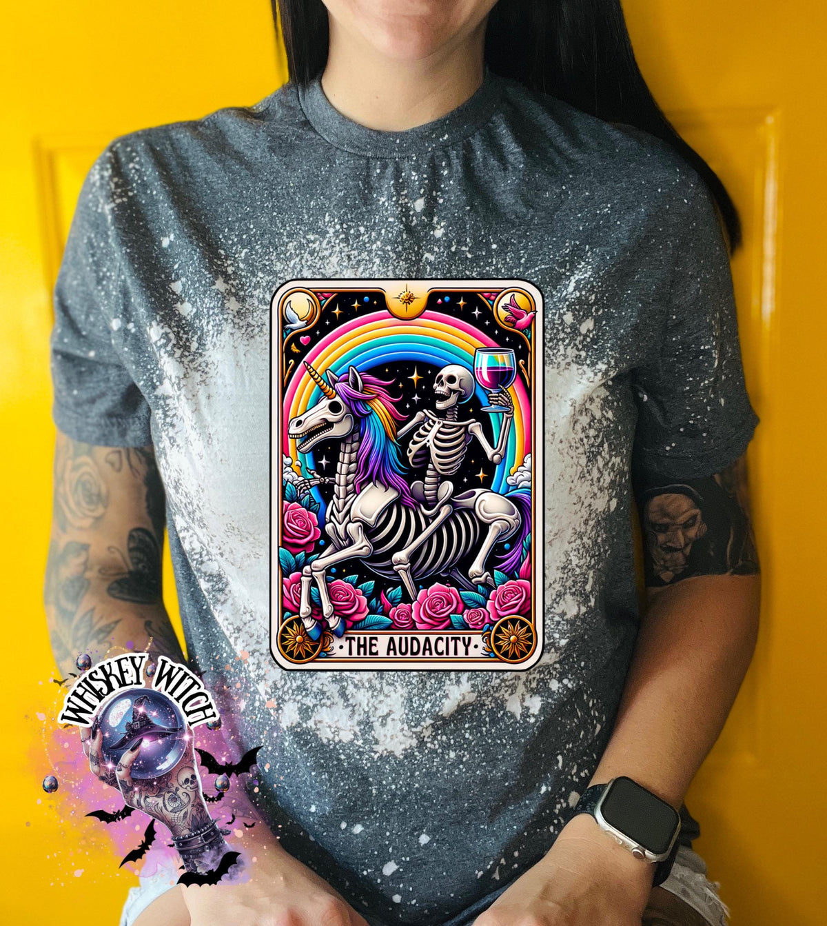 A grey  t-shirt with a bleached design featuring a tarot card with the image of  skeleton holding a glass of wine  riding a unicorn with a rainbow behind them with the words of “The Audacity” at the bottom