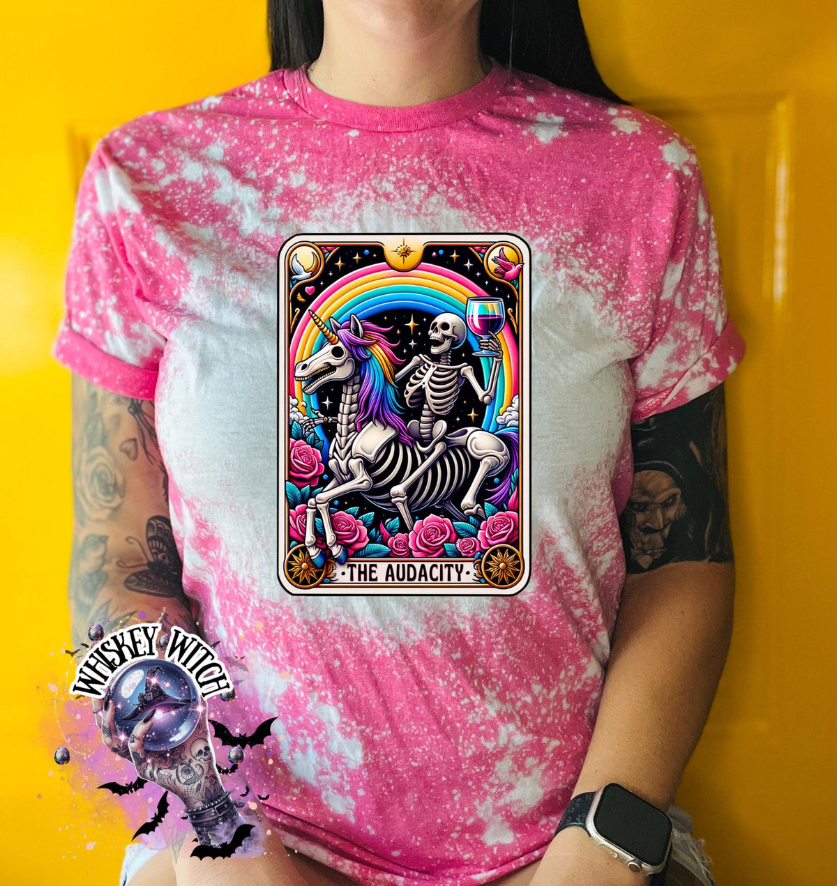 A pink  t-shirt with a bleached design featuring a tarot card with the image of  skeleton holding a glass of wine  riding a unicorn with a rainbow behind them with the words of “The Audacity” at the bottom