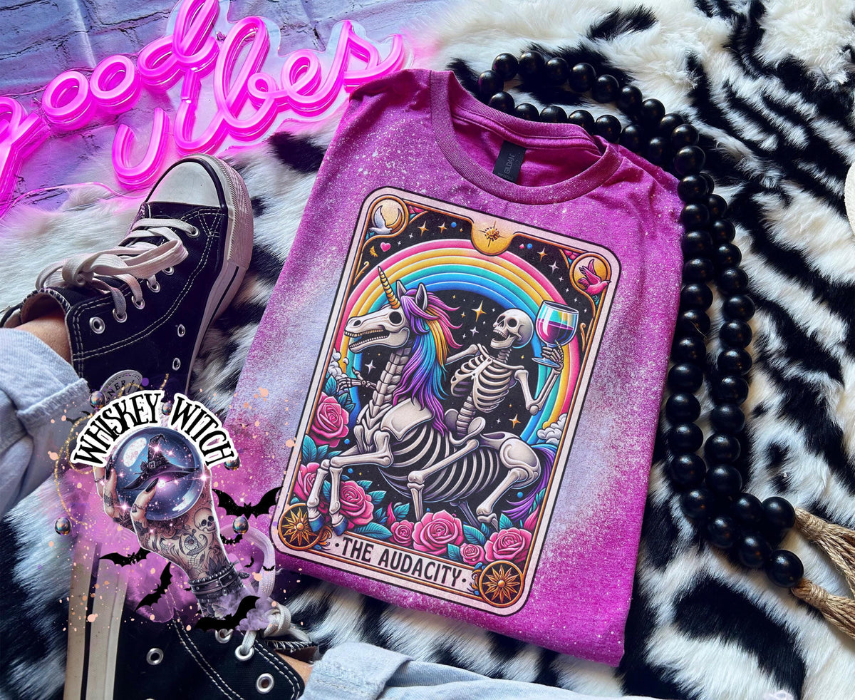 A berry pink  t-shirt with a bleached design featuring a tarot card with the image of  skeleton holding a glass of wine  riding a unicorn with a rainbow behind them with the words of “The Audacity” at the bottom