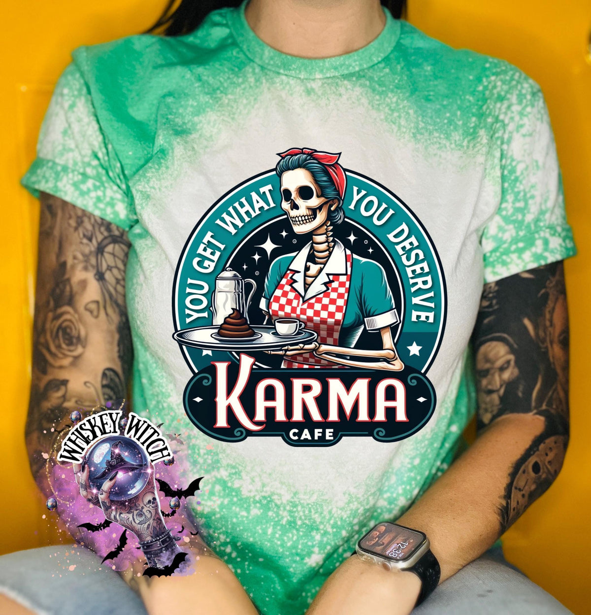 A green t-shirt with a bleached design featuring a skeleton wearing a red and white dress and holding a tray with a cup of coffee and a piece of cake. The text "You get what you deserve" is written around the skeleton. Below the skeleton is the text "Karma Cafe".