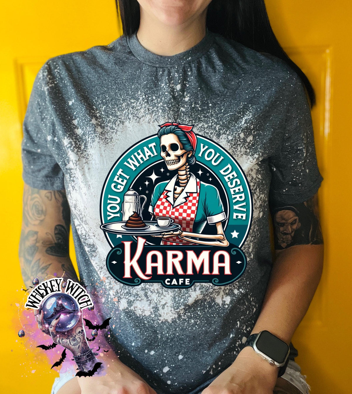 A grey t-shirt with a bleached design featuring a skeleton wearing a red and white dress and holding a tray with a cup of coffee and a piece of cake. The text "You get what you deserve" is written around the skeleton. Below the skeleton is the text "Karma Cafe".