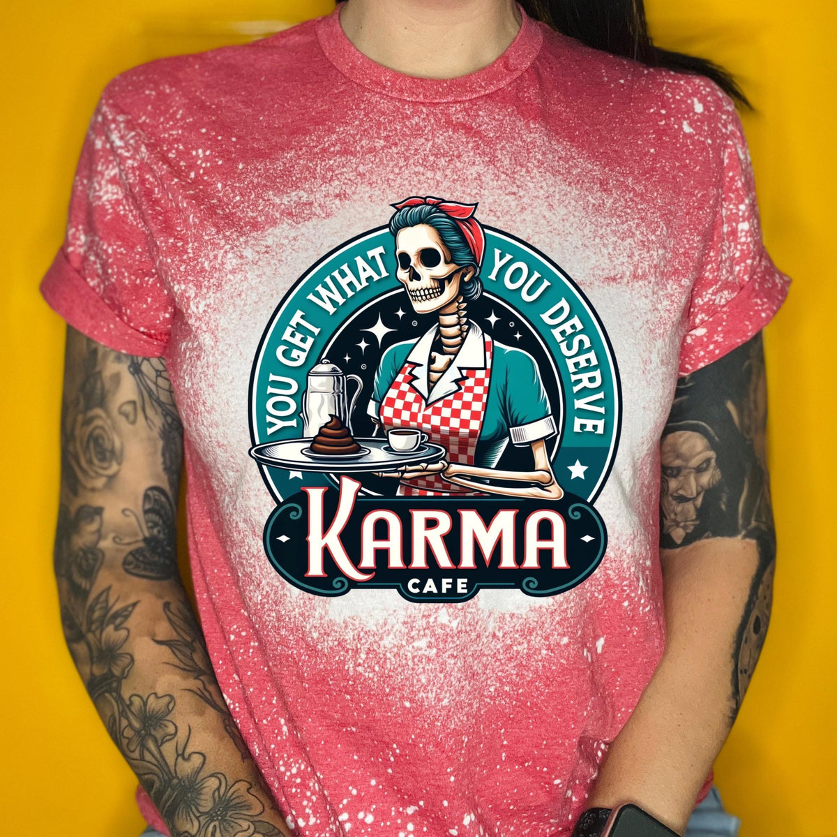 A red t-shirt with a bleached design featuring a skeleton wearing a red and white dress and holding a tray with a cup of coffee and a piece of cake. The text "You get what you deserve" is written around the skeleton. Below the skeleton is the text "Karma Cafe".