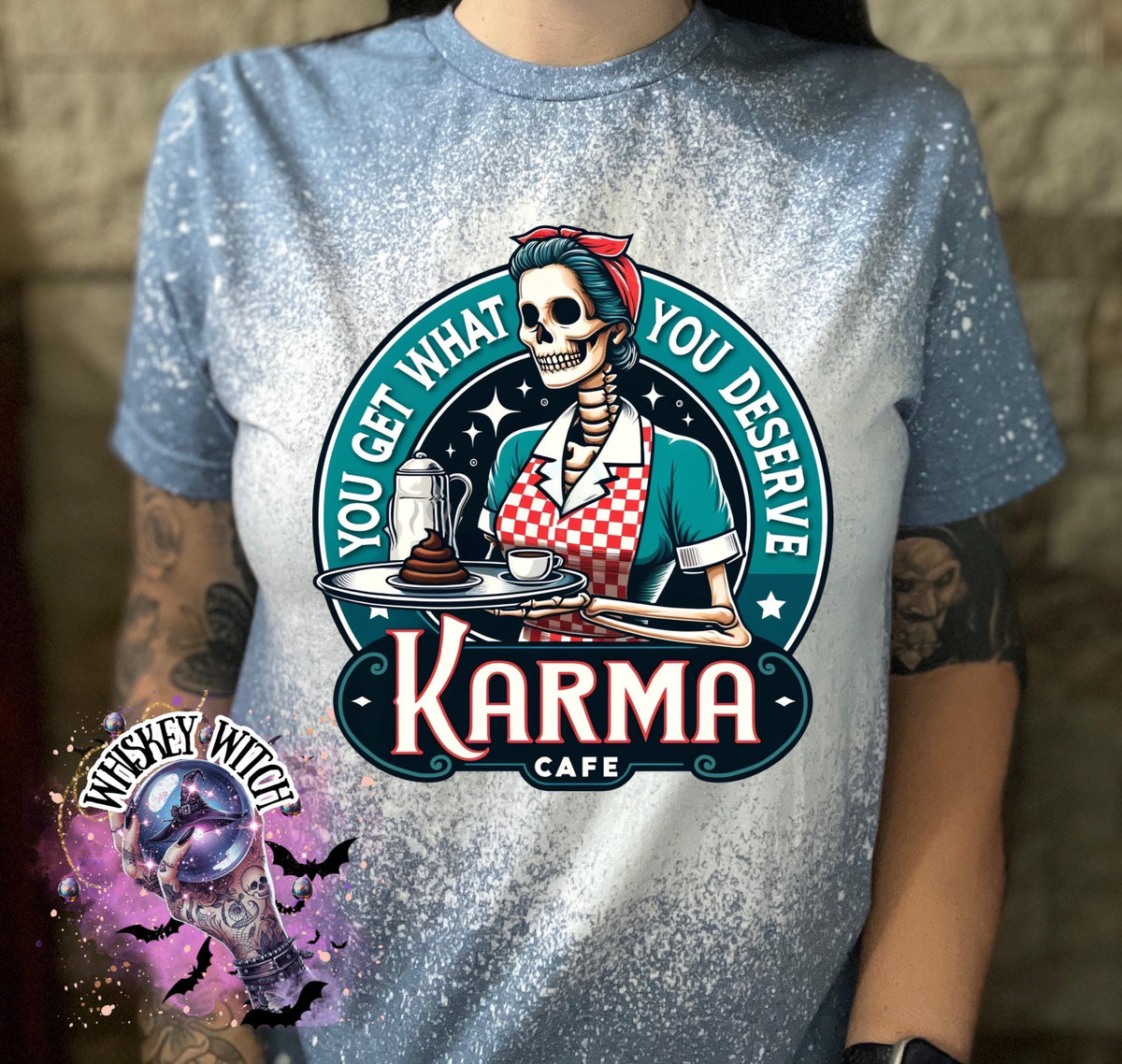 A indigo blue  t-shirt with a bleached design featuring a skeleton wearing a red and white dress and holding a tray with a cup of coffee and a piece of cake. The text "You get what you deserve" is written around the skeleton. Below the skeleton is the text "Karma Cafe".