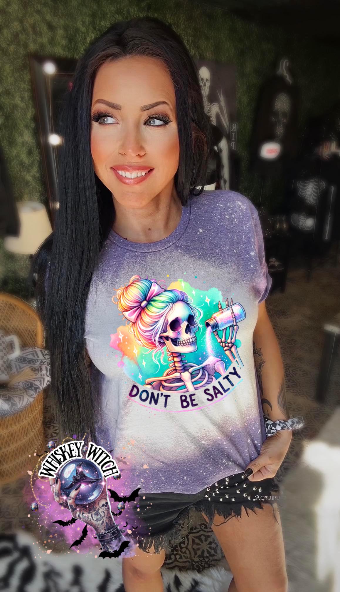 A purple  t-shirt with a bleached effect featuring a skeleton with rainbow hair pouring salt from a shaker. The text "Don't Be Salty" is printed on the shirt