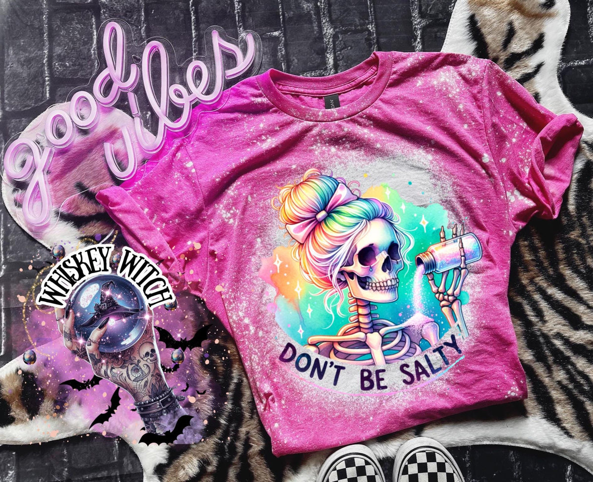 A pink  t-shirt with a bleached effect featuring a skeleton with rainbow hair pouring salt from a shaker. The text "Don't Be Salty" is printed on the shirt