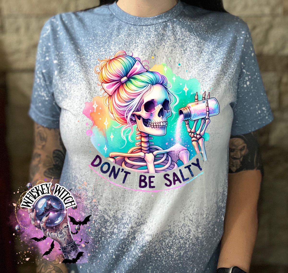 A indigo blue  t-shirt with a bleached effect featuring a skeleton with rainbow hair pouring salt from a shaker. The text "Don't Be Salty" is printed on the shirt