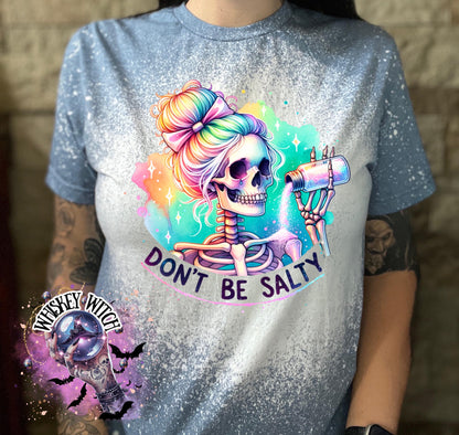 A indigo blue  t-shirt with a bleached effect featuring a skeleton with rainbow hair pouring salt from a shaker. The text "Don't Be Salty" is printed on the shirt