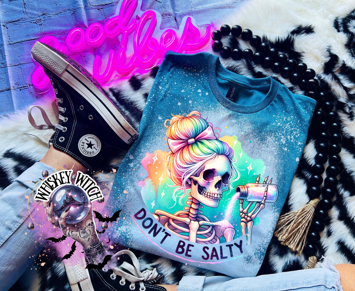 A Galapagos blue t-shirt with a bleached effect featuring a skeleton with rainbow hair pouring salt from a shaker. The text "Don't Be Salty" is printed on the shirt