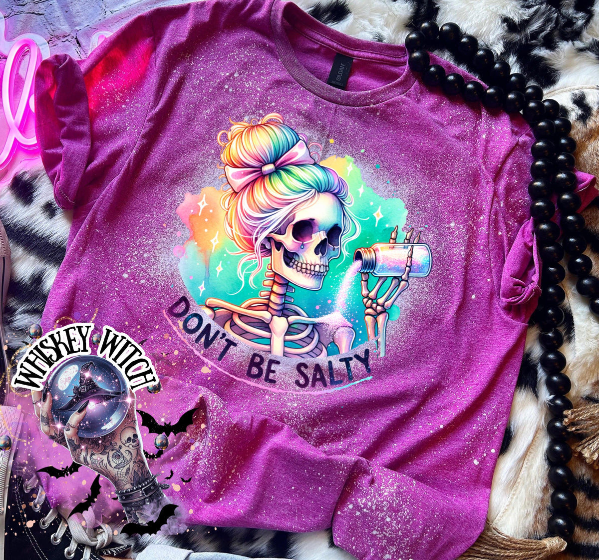 A Berry pink  t-shirt with a bleached effect featuring a skeleton with rainbow hair pouring salt from a shaker. The text "Don't Be Salty" is printed on the shirt