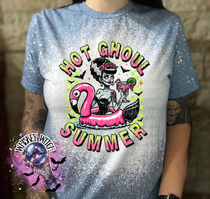 A indigo blue  and bleach splattered effect  t-shirt with a graphic of a zombie woman in a flamingo pool float, holding a cocktail. The text "Hot Ghoul Summer" is printed on the shirt.