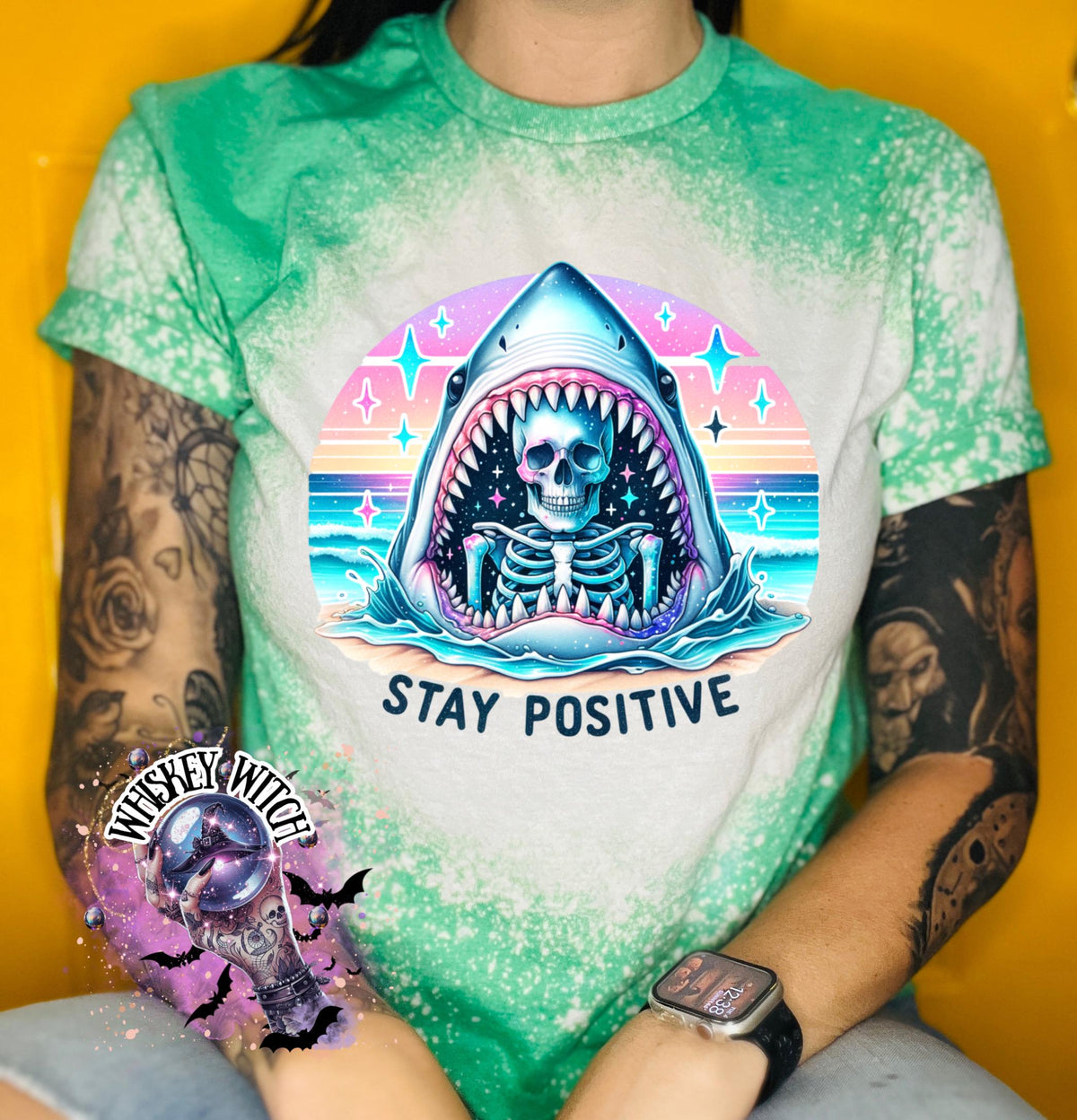 A green t-shirt with a bleached design of a skeleton inside a shark's mouth. The shark is surrounded by a rainbow and stars. The text "Stay Positive" is below the shark.