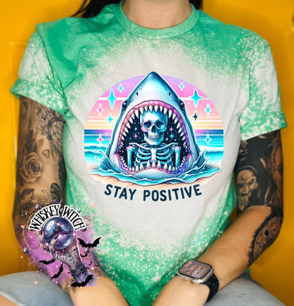 A green t-shirt with a bleached design of a skeleton inside a shark's mouth. The shark is surrounded by a rainbow and stars. The text "Stay Positive" is below the shark.