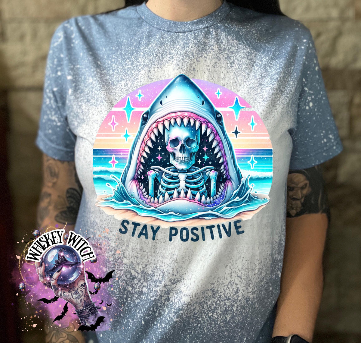 A indigo blue  t-shirt with a bleached design of a skeleton inside a shark's mouth. The shark is surrounded by a rainbow and stars. The text "Stay Positive" is below the shark.