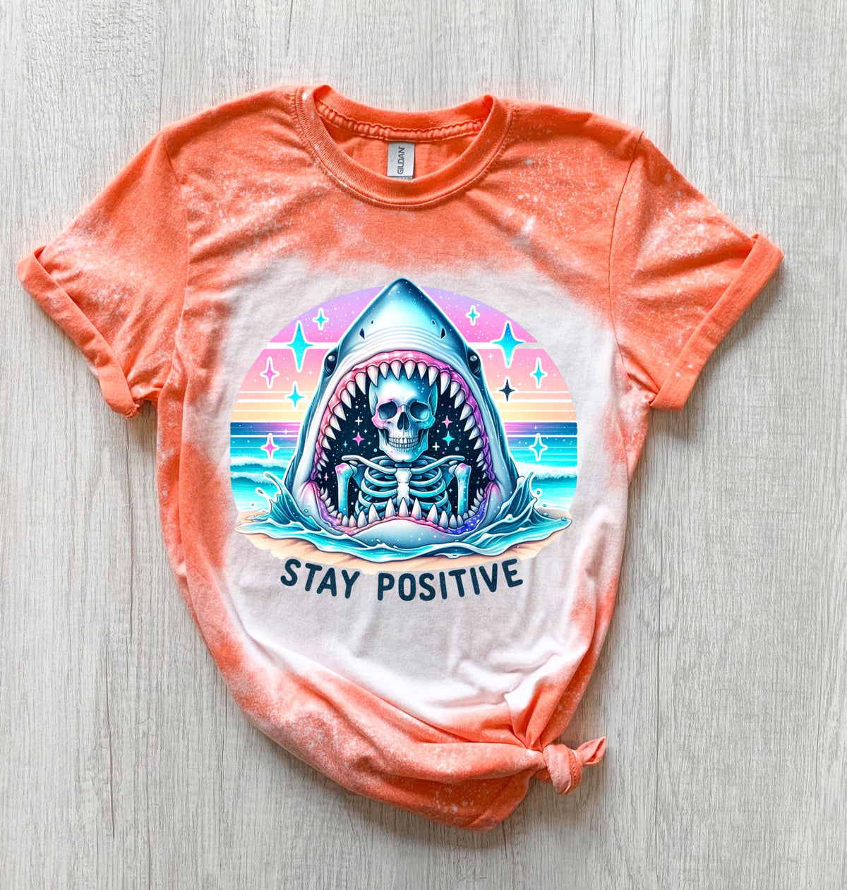 A Orange  t-shirt with a bleached design of a skeleton inside a shark's mouth. The shark is surrounded by a rainbow and stars. The text "Stay Positive" is below the shark.
