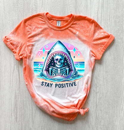 A Orange  t-shirt with a bleached design of a skeleton inside a shark's mouth. The shark is surrounded by a rainbow and stars. The text "Stay Positive" is below the shark.