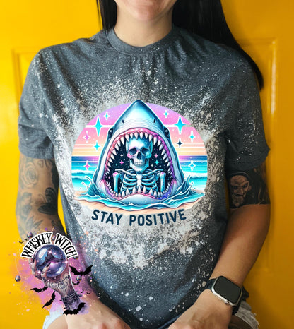 A gray t-shirt with a bleached design of a skeleton inside a shark's mouth. The shark is surrounded by a rainbow and stars. The text "Stay Positive" is below the shark.