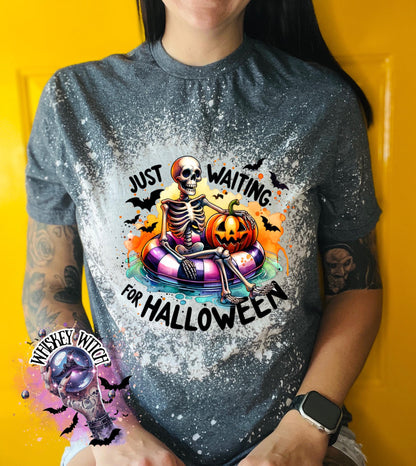 Dark Heather t-shirt with a bleached effect featuring a skeleton sitting in a pool float with a pumpkin. The text on the shirt reads "Just Waiting for Halloween".