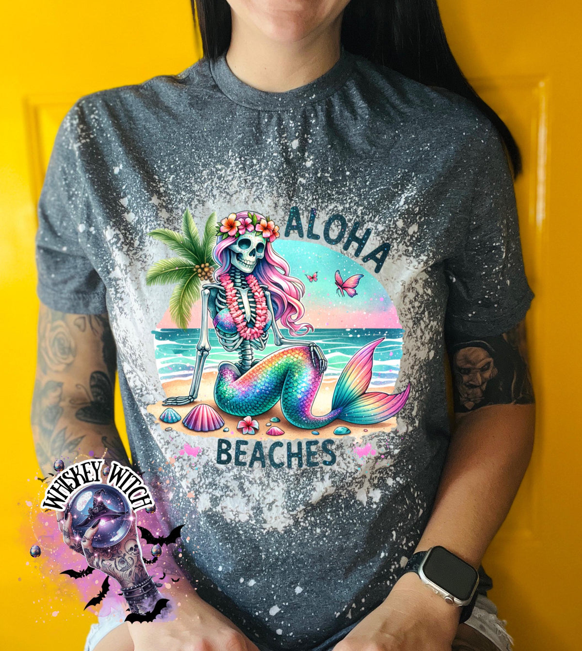 A grey t-shirt with a bleached effect features a colorful skeleton mermaid with a flower crown, palm tree, and seashells. The text "Aloha Beaches" is printed on the shirt.