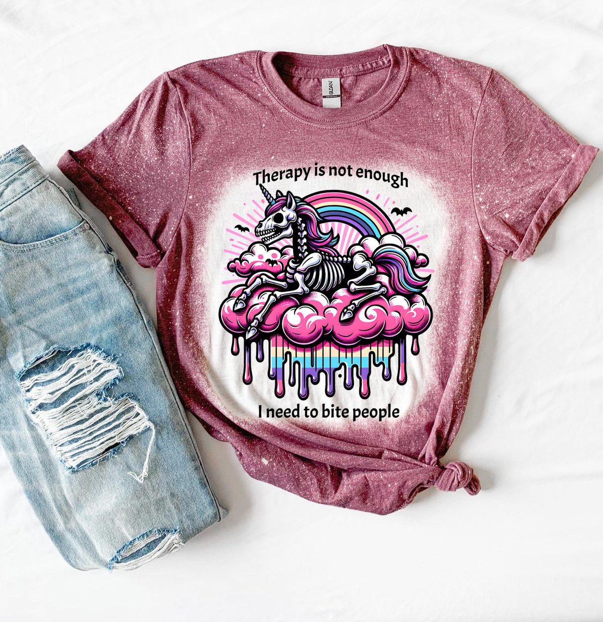 A maroon  and bleached design T shirt with a skeleton unicorn and a rainbow with a dripping colored cloud with the words “Therapy is not enough, I need to bite people.”