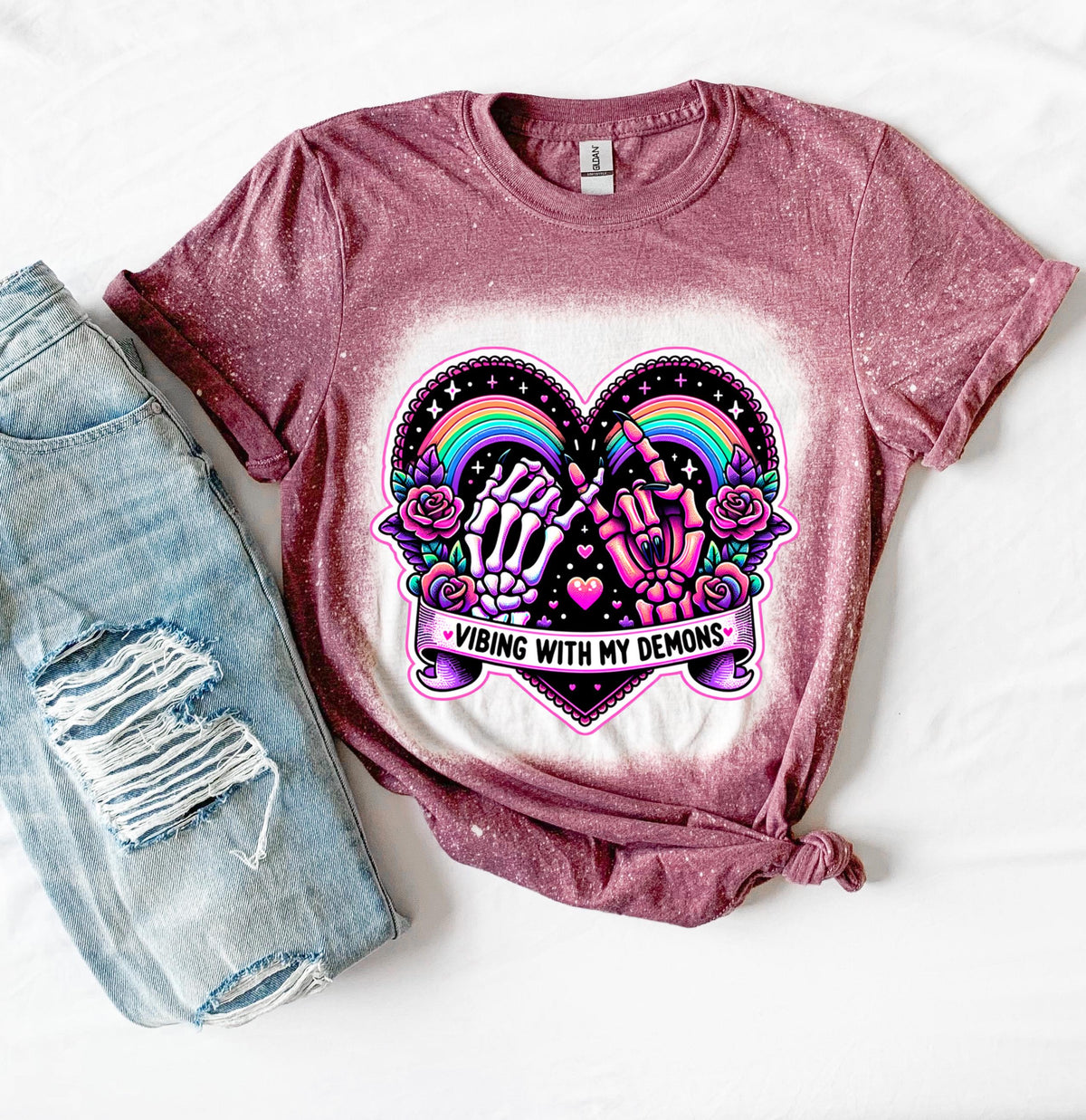 A maroon t-shirt with a bleach-splattered design featuring a pink heart with a rainbow and two hands, one skeleton and one with claws, holding pinky fingers together. The text 'Vibing with my demons' is written below the heart.
