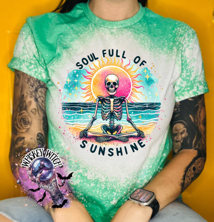 A green and bleach white splatter  t-shirt featuring a skeleton meditating on a beach with a sun in the background. The text on the shirt reads "SOUL FULL OF SUNSHINE".