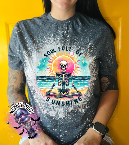 Dark gray t-shirt with bleach splatter with  a colorful graphic of a skeleton meditating in front of a sun and ocean scene. The text "SOUL FULL OF SUNSHINE" is printed below the image. The shirt has a bleached, distressed look.