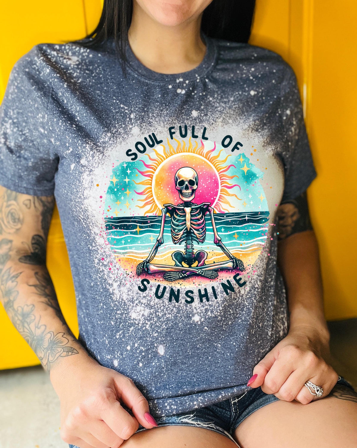 A naby blue and bleach white splatter  t-shirt featuring a skeleton meditating on a beach with a sun in the background. The text on the shirt reads "SOUL FULL OF SUNSHINE".