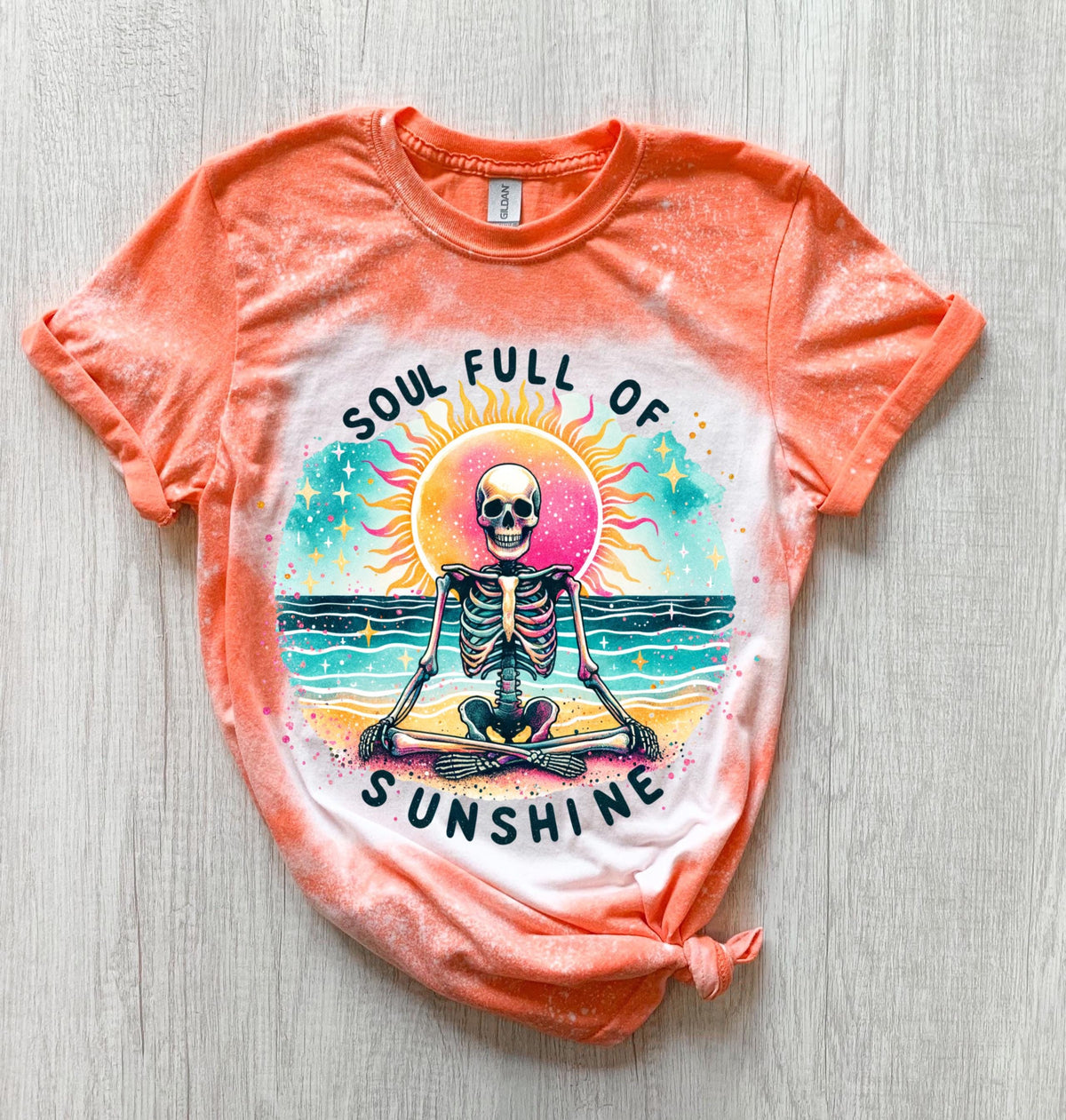 A Orange and bleach white splatter  t-shirt featuring a skeleton meditating on a beach with a sun in the background. The text on the shirt reads "SOUL FULL OF SUNSHINE".