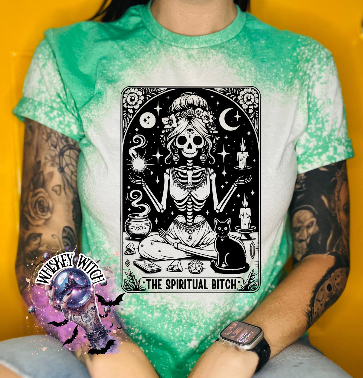 A bleached green t-shirt with a black and white illustration of a skeleton woman with a cat and the text "The Spiritual Bitch".