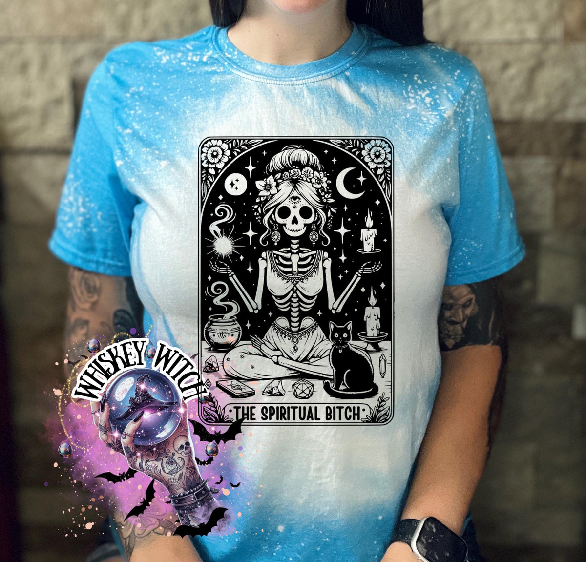 A bleached saphire blue t-shirt with a black and white illustration of a skeleton woman with a cat and the text "The Spiritual Bitch".