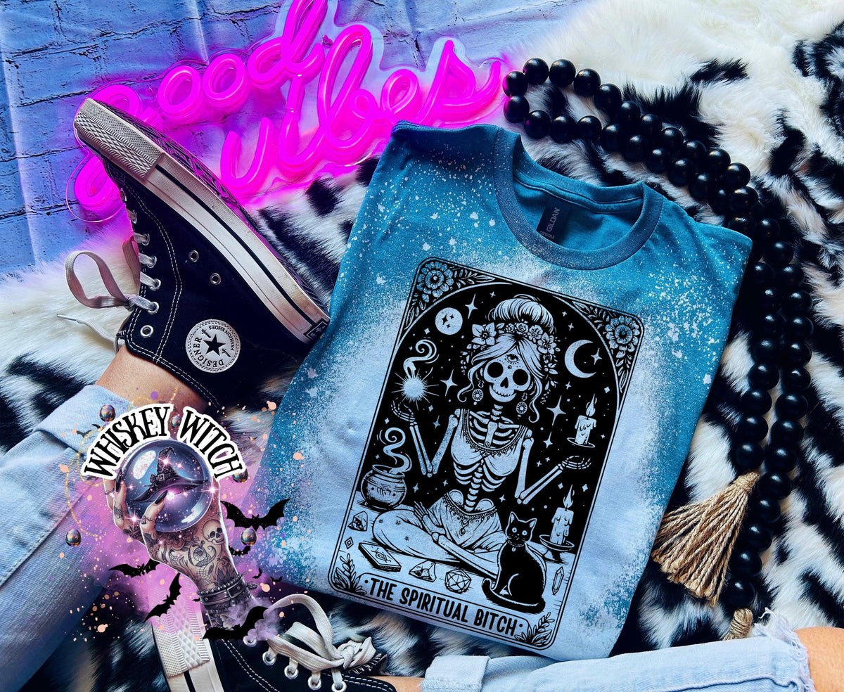 A bleached Galapagos blue t-shirt with a black and white illustration of a skeleton woman with a cat and the text "The Spiritual Bitch".