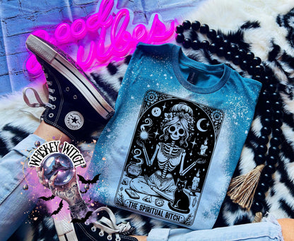A bleached Galapagos blue t-shirt with a black and white illustration of a skeleton woman with a cat and the text "The Spiritual Bitch".