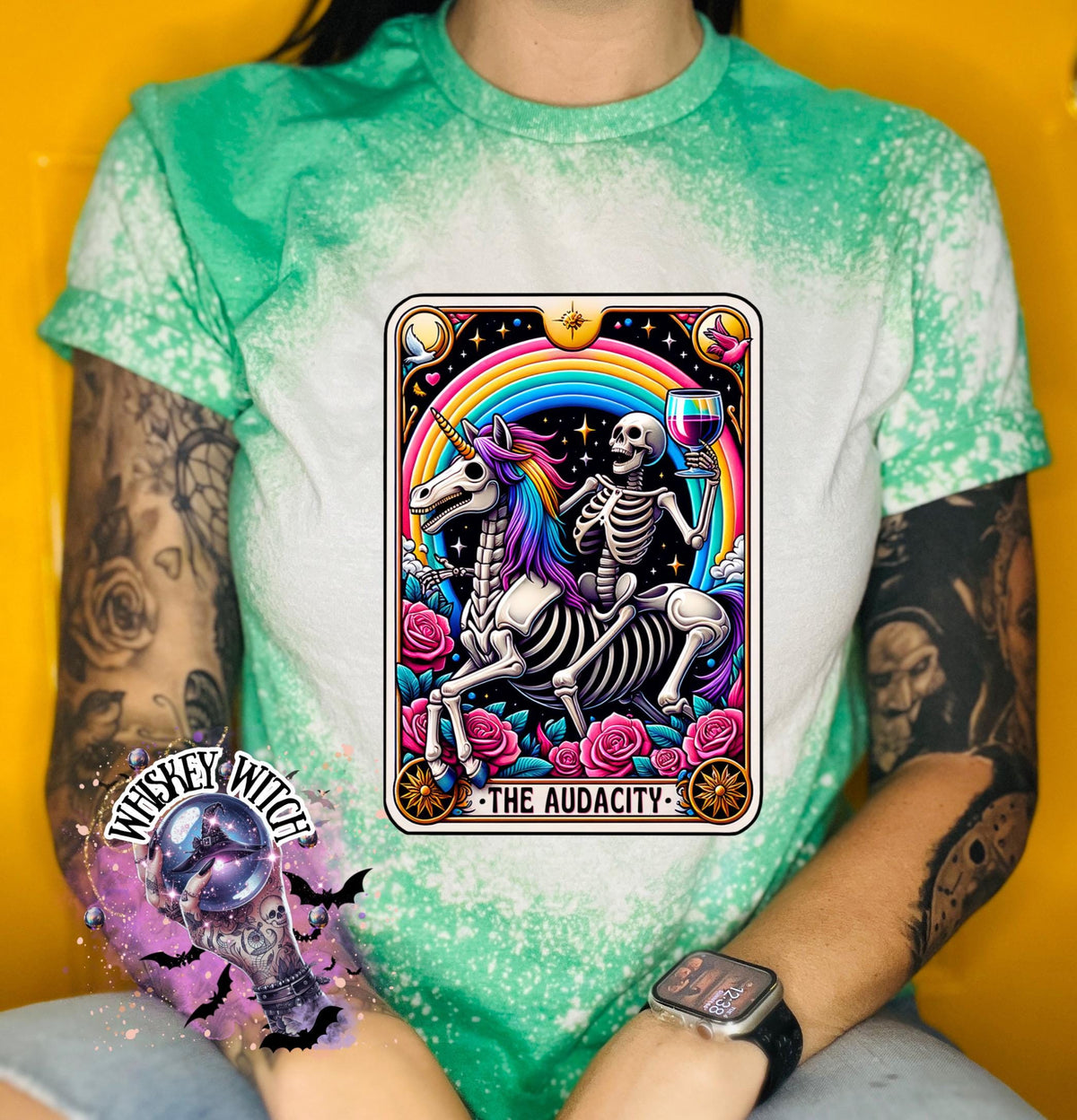 A green  t-shirt with a bleached design featuring a tarot card with the image of  skeleton holding a glass of wine  riding a unicorn with a rainbow behind them with the words of “The Audacity” at the bottom