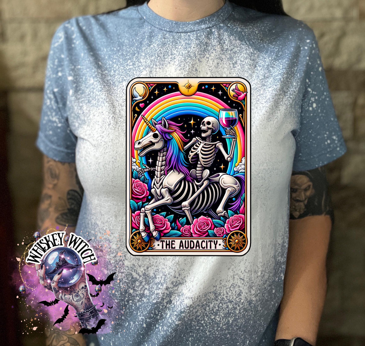 A indigo blue  t-shirt with a bleached design featuring a tarot card with the image of  skeleton holding a glass of wine  riding a unicorn with a rainbow behind them with the words of “The Audacity” at the bottom